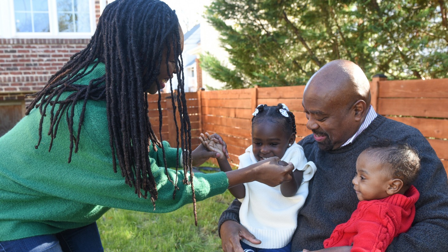在一个阳光明媚的后院，一位黑人妈妈与她的小女儿握手，她的小女儿正与哥哥坐在父亲的腿上。
